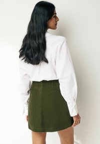 Femeie cu păr lung și negru, purtând o cămașă albă cu mânecă lungă și o fustă din bumbac de culoare verde măsliniu, stând lateral lângă un zid alb.