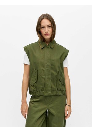Femme à la peau claire et aux cheveux bruns portant une veste sans manches vert olive sur une chemise blanche à manches courtes et un pantalon vert assorti.