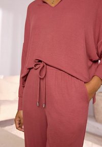 Knit top v rezavé barvě s výstřihem do V a volnými rukávy, v kombinaci s odpovídajícími kalhotami s nastavitelným pasem a jemnou texturou.