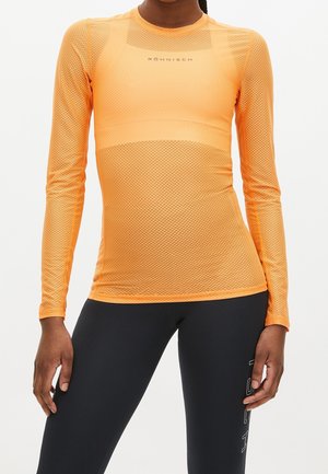 Osoba nosząca pomarańczową, siateczkową bluzkę z długim rękawem oraz czarne legginsy z białym napisem logo na udzie, stojąca na jednolitym tle.