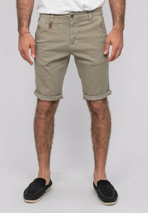 Homme portant un short beige retroussé, des chaussures noires sans lacets et une chemise blanche, debout devant un fond clair uni.