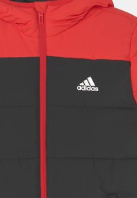 Veste matelassée avec une capuche et une partie supérieure rouges, une partie inférieure noire, une fermeture éclair à l'avant, et un logo Adidas blanc sur le côté gauche de la poitrine.