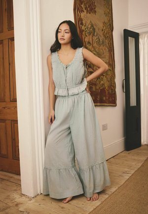 Woman with long dark hair stands barefoot in a room, wearing light blue sleeveless top and wide-leg pants with ruffled hems.