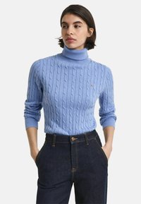 Lichtblauwe kabelgebreide coltrui met geribde manchetten, gedragen met donkere denim broek met contrasterende stiksels en een voorknoop.