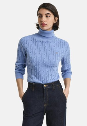 Lichtblauwe kabelgebreide coltrui met geribde manchetten, gedragen met donkere denim broek met contrasterende stiksels en een voorknoop.