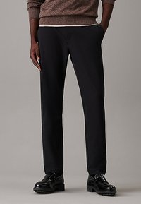 Pantaloni neri su misura con una vestibilità liscia e tagliata, dotati di tasche laterali e un design frontale piatto. Indossati con scarpe nere su uno sfondo neutro.