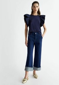 Top blu navy con grandi maniche a volant in raso, abbinato a jeans a gamba larga blu scuro con orlo risvoltato. Gli accessori includono scarpe con tacco slingback dorate.
