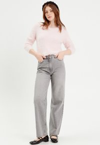 Lichtroze fuzzy sweater, hooggepaste grijze jeans en zwarte lakleren ballerina's. Lange mouwen met ribgebreide manchetten. Model poseert met handen op de heupen.
