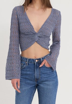 Femme portant un crop top en tricot lavande à manches longues avec un twist à l'avant et un jean bleu taille haute, main dans la poche.