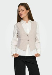 Gilet en laine beige clair avec un col en V, deux boutons à l'avant et une coupe ajustée, porté sur une chemise blanche à col avec des accents en perles.