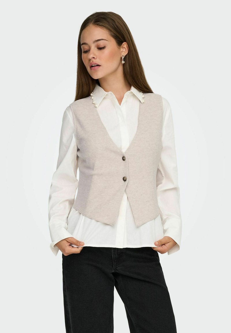 Gilet en laine beige clair avec un col en V, deux boutons à l'avant et une coupe ajustée, porté sur une chemise blanche à col avec des accents en perles.