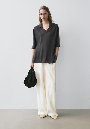 Kvinde står op ad en ensfarvet væg iført en løs, mørk overdel, brede hvide bukser og holder en sort taske i den ene hånd, beige sko er synlige.