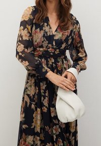 Woman wearing a long floral dress with sheer sleeves, holding a folded white handbag in front of a plain background.