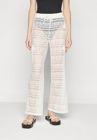 Pantalons en crochet blanc avec un motif en zigzag et des jambes évasées. Ceinture élastique avec cordon de serrage. Portés avec des sandales noires plates.
