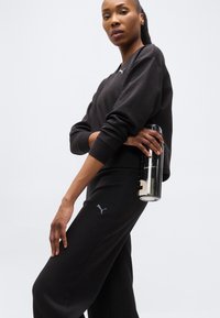 Γυναίκα με μαύρη φόρμα Puma, κρατώντας ένα μαύρο παγούρι νερού, μπροστά σε απλό φωτεινό φόντο, κοιτώντας την κάμερα με αυτοπεποίθηση.