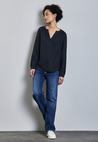 Blusa negra de manga larga con escote en V y mangas ligeramente fruncidas, combinada con jeans rectos azules y zapatillas blancas.