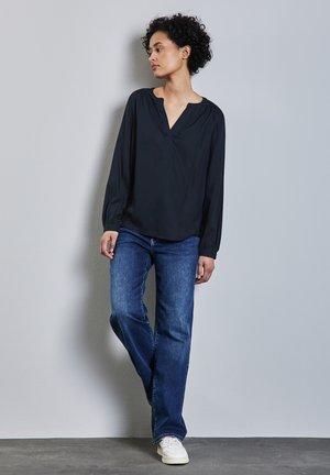 Blusa negra de manga larga con escote en V y mangas ligeramente fruncidas, combinada con jeans rectos azules y zapatillas blancas.