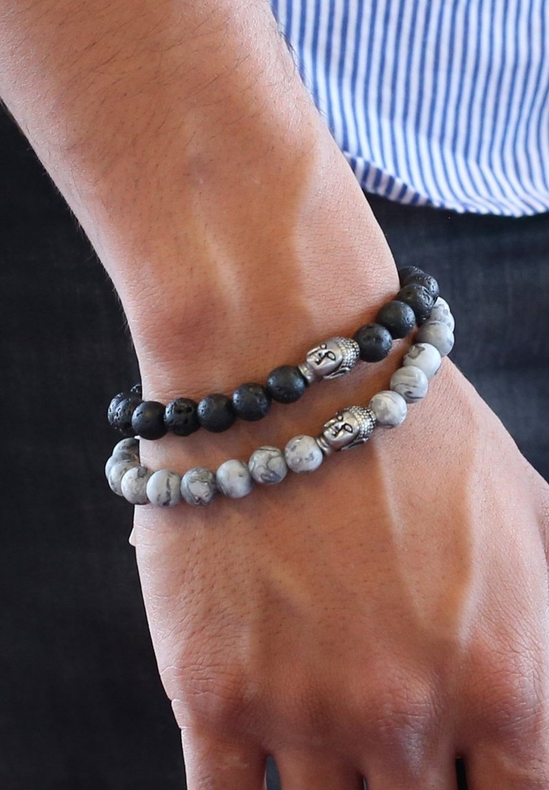 Twee kralenarmbanden om een pols: de ene van zwart lavasteen met ronde kralen, de andere van lichtgrijze marmer met een boeddha-hoofd accent.