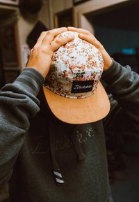 Floral-patterned cap with a beige suede brim, featuring a black label with "Blackskies," held by a person wearing a gray hoodie.