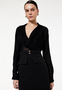 Blusa nera avvolgente con scollatura a V profonda, maniche lunghe e colletto. Abbinata a una gonna nera a vita alta con tasche applicate e una cintura con fibbia dorata.