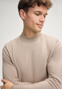 Beige, leichter Pullover mit langen Ärmeln, hohem Kragen und gerippten Bündchen. Glatte Textur, figurbetonte Passform, minimale Nähdetails.