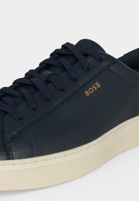 Gros plan d'une basket en cuir bleu marine avec des lacets foncés et une semelle blanche, portant le texte de la marque "BOSS" en lettres dorées sur le côté.