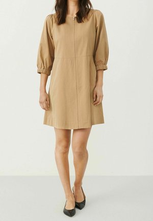 Femme portant une robe beige mi-longue aux manches bouffantes trois-quarts et une fermeture éclair devant, associée à des escarpins noirs à bout pointu, debout contre un mur blanc.