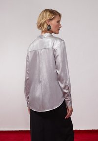 Sølvfarget, strukturert bluse med lange ermer og krage, kombinert med et svart skjørt. Har knapper på ermene.