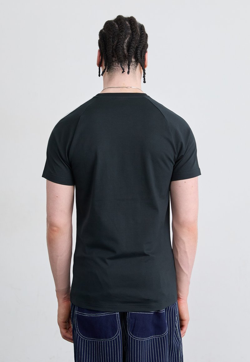 Schwarzes T-Shirt mit kurzen Ärmeln, glatter Textur und Raglanärmeln, von hinten gesehen. Kombiniert mit blauen vertikal gestreiften Hosen.