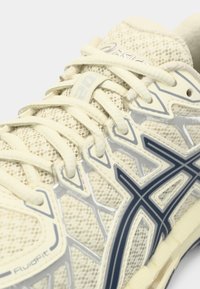 Beige αθλητικά παπούτσια Asics για τρέξιμο με πλέγμα στην επάνω επιφάνεια, κρέμα κορδόνια και μαύρη λεπτομέρεια λογότυπου στο πλάι, με κοντινή λήψη της περιοχής των κορδονιών.
