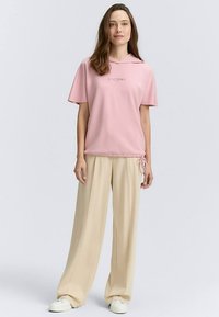 Roosa lühikeste varrukatega kapuutsiga särk tikitud tekstiga, kombineeritud helebeežide laiade pükstega ja valgete tossudega.