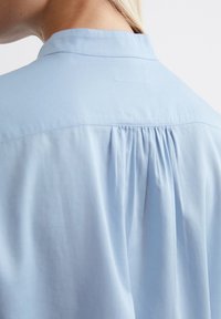 Light blue shirt with a collar, featuring a gathered texture at the back, made from a smooth fabric, showing soft, flowing lines.