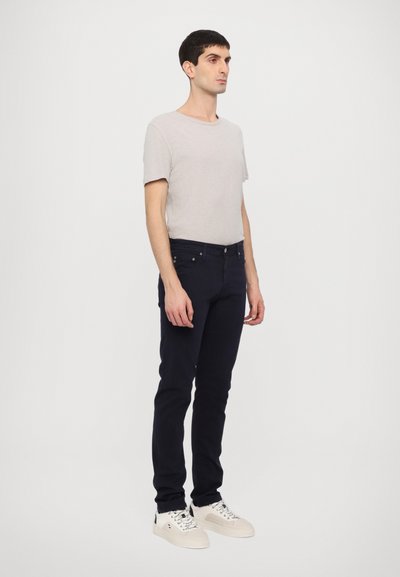 Ανοιχτό γκρι κοντομάνικο μπλουζάκι, ναυτικές τζιν και λευκά αθλητικά παπούτσια. Τα τζιν είναι εφαρμοστά με μπροστινές τσέπες και κλείσιμο με φερμουάρ.