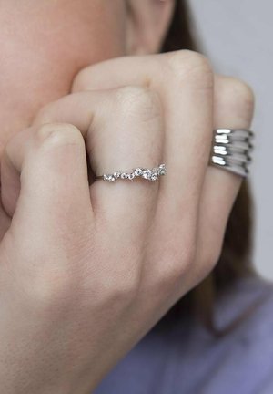 Anillo de plata con diseño de ola, adornado con pequeñas piedras preciosas transparentes dispuestas de manera alterna, exhibido en una mano con una textura de piel suave y clara.