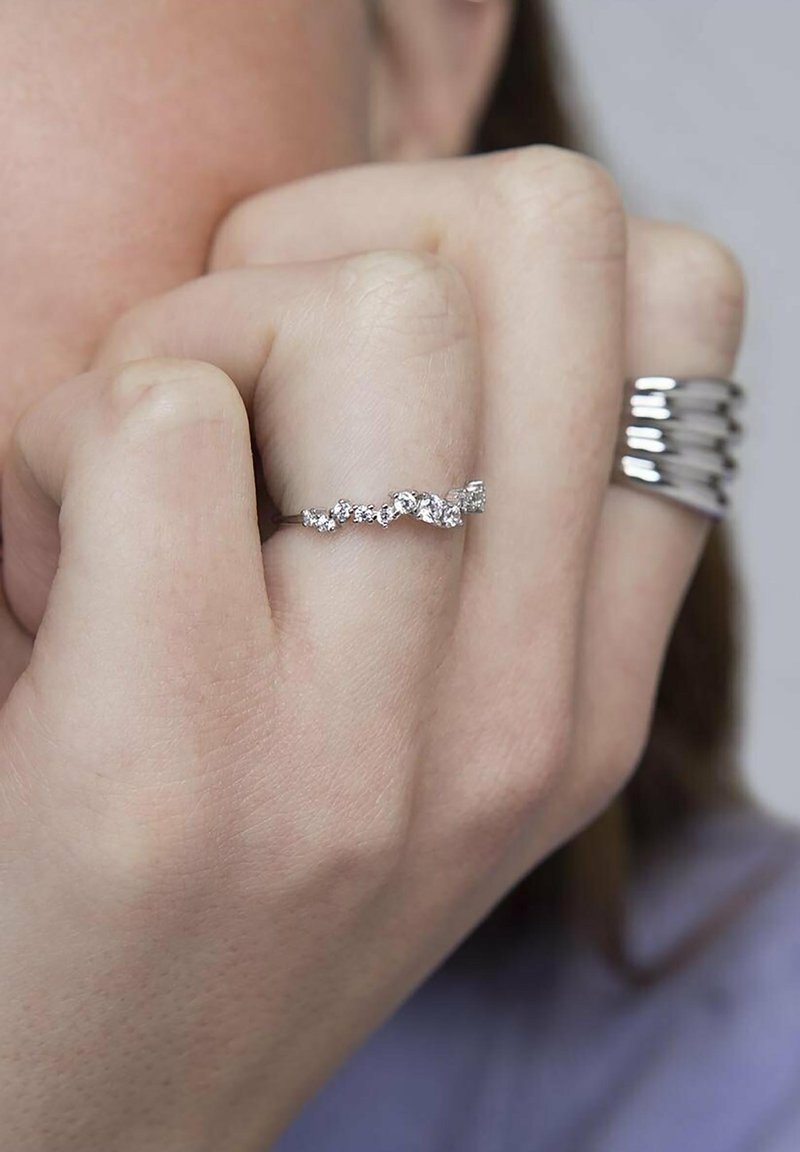 Anello in argento con design a onda, dotato di piccoli gemme trasparenti incastonate alternativamente, esposto su una mano con una texture della pelle liscia e chiara.