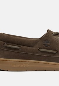 Brauner Wildleder-Bootsschuh mit grünen Akzenten, runden Ösen und einer Gummisohle. Timberland-Logo ist seitlich und auf der Sohle geprägt.
