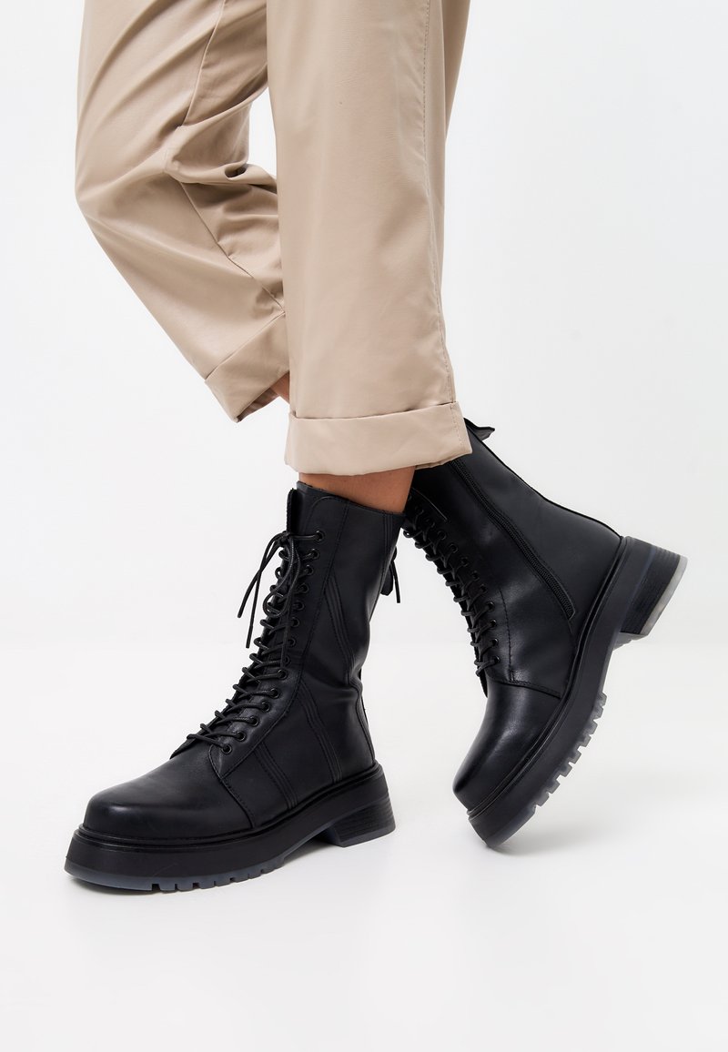 Botas de combate de cuero negro con cordones, puntera redonda y entresuela texturizada. Combinadas con pantalones beiges con puños.