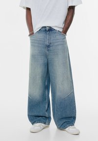 Široke modre kavbojke z zbledelimi področji, opremljene z gumbom in zadrgo, dvema sprednjima žepoma ter šivanjem na hlačnicah.