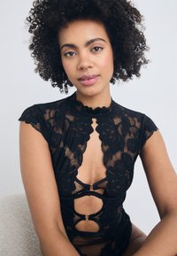 Black lace bodysuit with floral pattern, high collar, keyhole cutouts, and gold accents. Short sleeves and sheer texture.