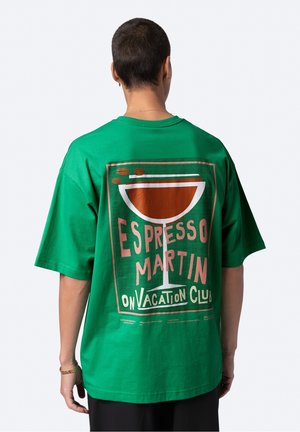 Person mit kurzen Haaren trägt ein grünes, übergroßes T-Shirt mit einem Grafikdruck eines Espresso-Martini-Cocktails und Text auf der Rückseite.