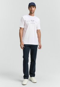Hvid kortærmet t-shirt med marineblå grafisk tryk, parret med marineblå bukser og hvide sneakers, sat mod en neutral baggrund.