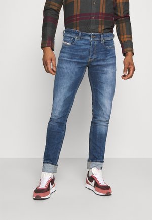 Homme portant un jean bleu retroussé, des baskets rouges et blanches, et une chemise à manches longues à carreaux, debout devant un fond uni.