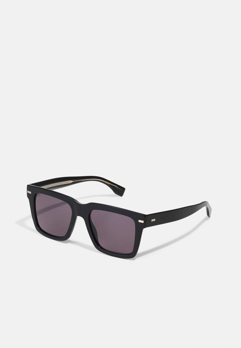Lunettes de soleil carrées noires avec des verres teintés violets. Présentent de fines accents dorés sur les côtés de la monture. Fabriquées en plastique brillant.
