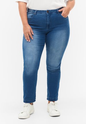 Blaue Slim-Fit-Jeans aus Denim mit einem ausgewaschenen Finish, ausgestattet mit fünf Taschen und einem einzigen Knopfverschluss vorne. Kombiniert mit weißen Sneakers.