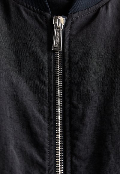 Close-up van een zwarte jasrits met metalen tanden en een rechthoekige ritstrekkerschijf gegraveerd met "DSQUARED2."