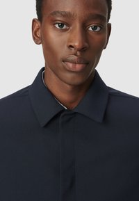 Chaqueta azul marino con un collar estructurado, cierre de botones y tela texturizada. Presentada en la parte superior del torso de un modelo, frente a un fondo liso.