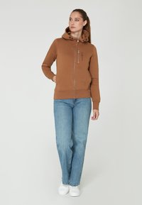 Sudadera con cremallera marrón, con capucha ajustable, bolsillos tipo canguro y logo bordado. Combinada con jeans de color azul claro y zapatillas blancas.