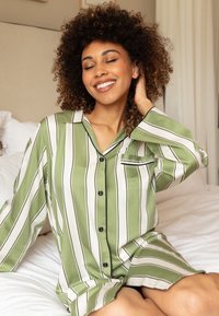 Zielono-biała pasiaste piżama z czarnymi akcentami, z kieszonką, luźny krój i gładki materiał, noszona w swobodny sposób na jasnym tle.