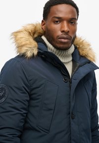 Parka blu navy con cappuccio in finta pelliccia, chiusura a bottoni e zip anteriore. Presenta un maglione a collo alto a coste sotto e una toppa sulla manica.