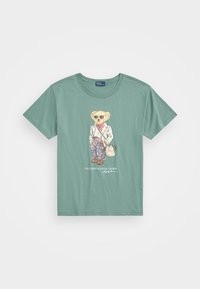 Camiseta de algodón verde con un oso de dibujos animados con gafas de sol, vistiendo un blazer blanco, pantalones a cuadros y sosteniendo una bolsa. Diseño gráfico en la parte delantera.